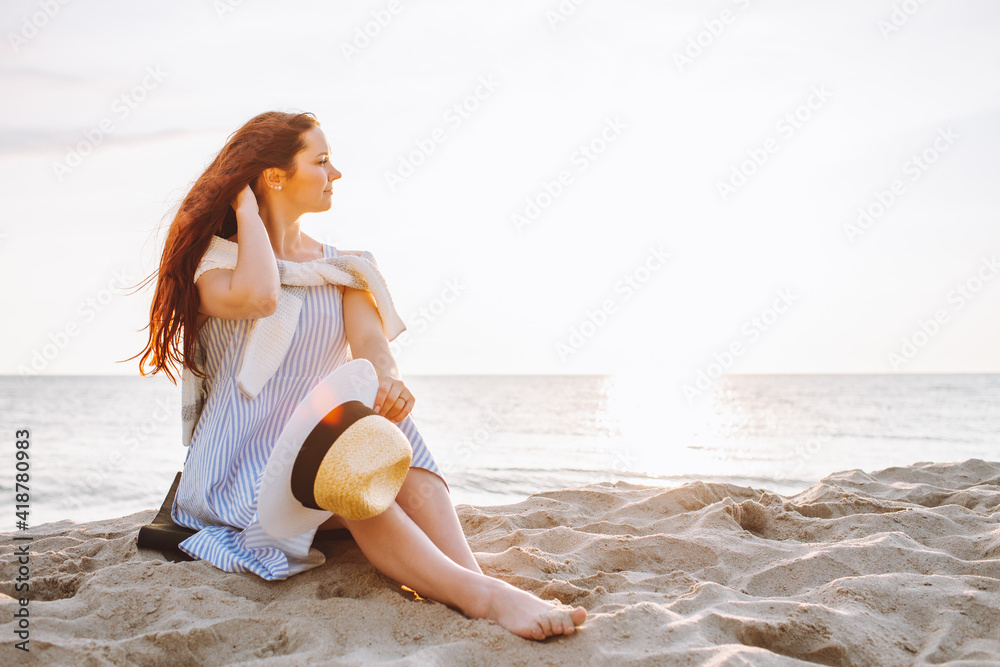 Naklejka premium Young woman in dress and straw hat sit alone on empty sand beach at summer sunset on sea shore and and smile. Space for message.