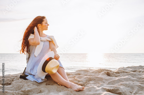 Young woman in dress and straw hat sit alone on empty sand beach at summer sunset on sea shore and and smile. Space for message.