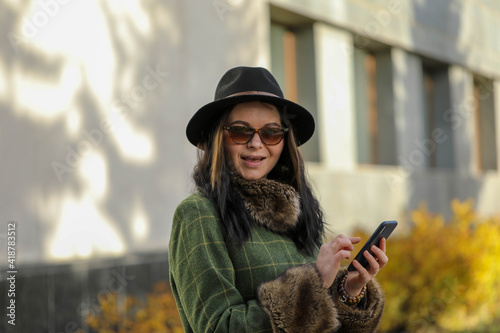 Beautiful woman in coat and hat uses smartphone while walking in spring city. She takes a selfie or makes a video call .