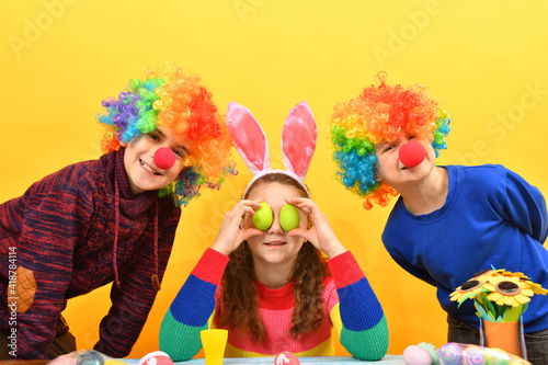 Girl holding Easter eggs in front of her eyes two clown boys are standing side by side.