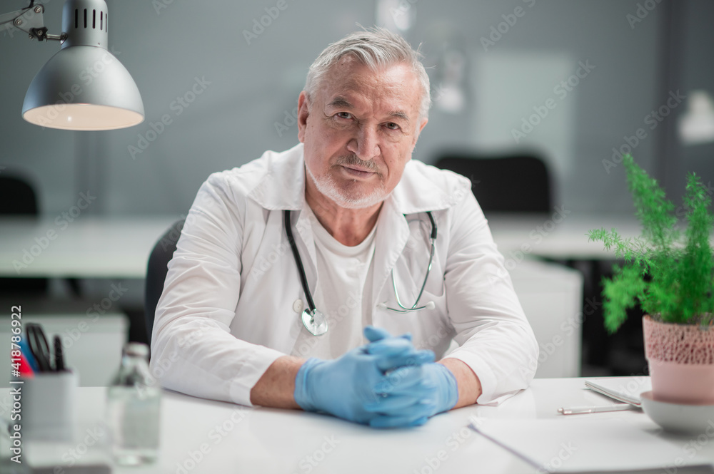 Portrait of a gray-haired doctor, who sits at a desk in the office in a dressing gown and with a stethoscope. Portrait of a gray-haired doctor, who sits at a desk in the office in a dressing gown and with a stethoscope.