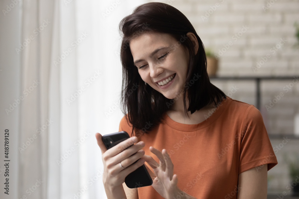 Happy teenage female stand at living room work on smartphone gadget online use wifi internet. Laughing young woman touch screen of modern phone device chat having fun scroll web pages read funny news Happy teenage female stand at living room work on smartphone gadget online use wifi internet. Laughing young woman touch screen of modern phone device chat having fun scroll web pages read funny news