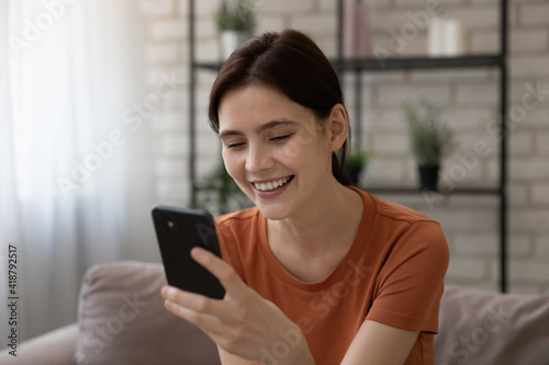 Happy smiling young female sit on sofa hold mobile phone read love message in personal chat with beloved man enjoy positive news in app. Glad teenager use cell gadget texting with good friend online
