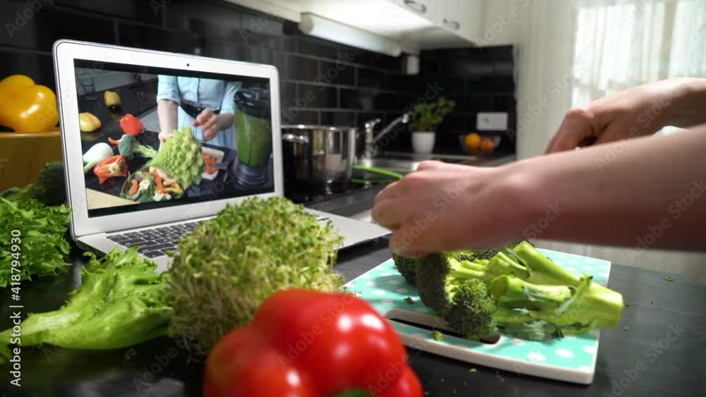 A young woman watching a online stream broadcast video blog tutorial about tasty and healthy food using laptop and cooking a meal at the home kitchen