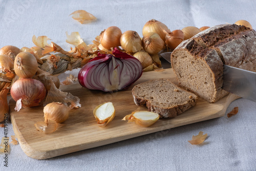 On a linen tablecloth, an ash cutting board with small whole and chopped golden onions and a half of a large red onion, fresh black bread, from which a large chunk is cut with a knife. 