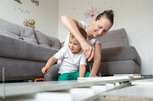 Obraz na plátne Cute adorable caucasian toddler boy kid sit on floor and help mom assembling furniture shelf with power screwdriver tool