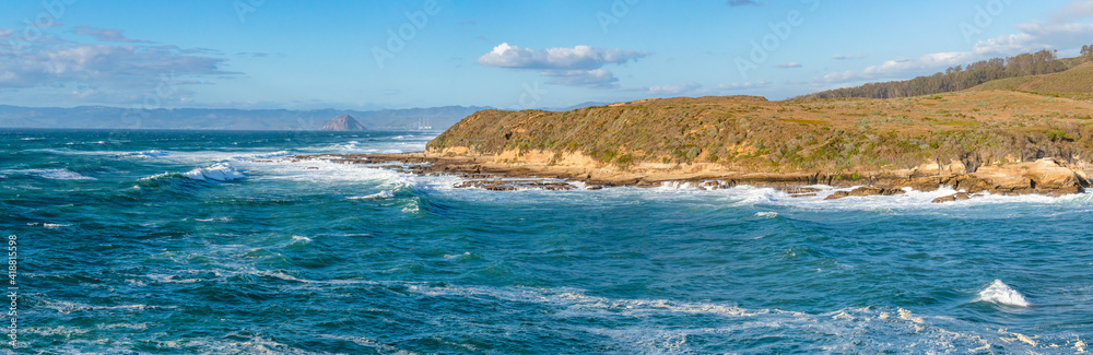 Panoramic seascape in blue and turquoise colors. Pacific ocean, rocky cliffs, and silhouette of Morro Rock. View from Montana del Oro state park, California coastline