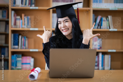 A young Asian female university graduate expressing joy and excitement to celebrate her achievement of degree graduation in front of a laptop making a remote video call to her parents at home