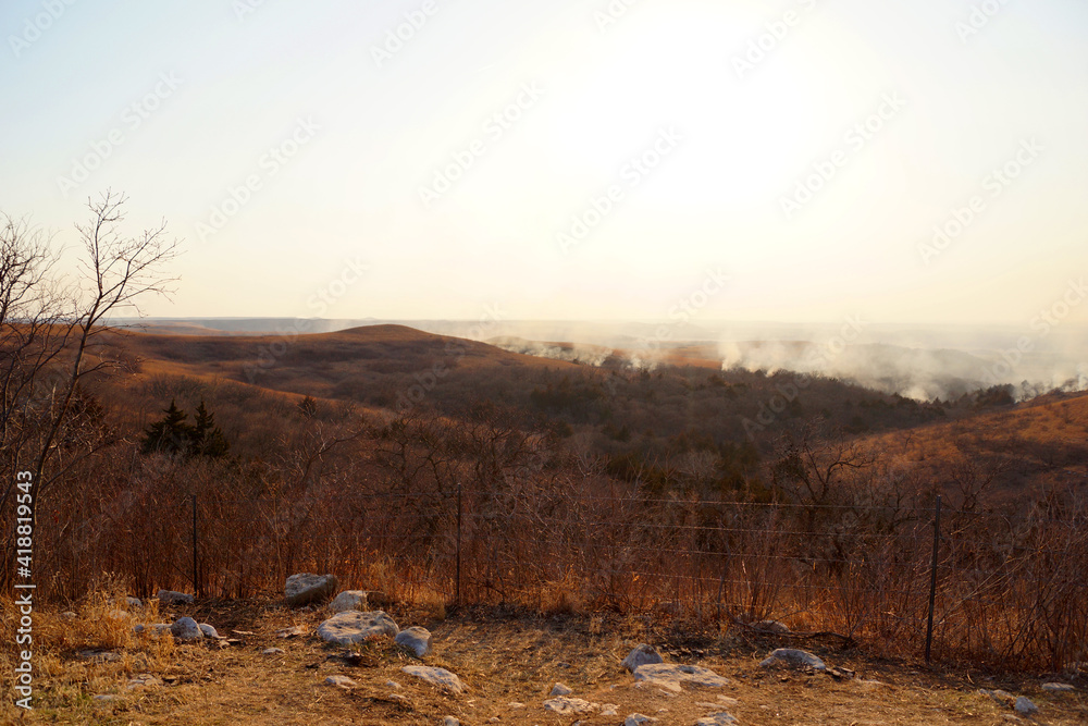 Wildfires in the distance burn brush and dry grass in the hills and fills the valley with smoke. A barbed wire fence in the foreground separates pastures on a ranch. Wildfires in the distance burn brush and dry grass in the hills and fills the valley with smoke. A barbed wire fence in the foreground separates pastures on a ranch.