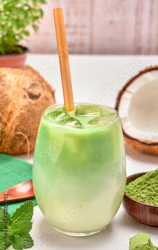 organic green tea latte with milk and a bamboo straw. on the side a wooden bowl with matcha powder, mint leaves and a couple of coconuts with a background of white wooden boards. copy space