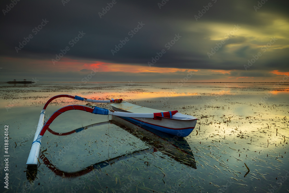 Fototapeta premium Seascape. Fisherman boat. Traditional Balinese boat jukung. Fishing boat at the beach during sunrise. Water reflection. Cloudy sky. Slow shutter speed. Soft focus. Sanur beach, Bali