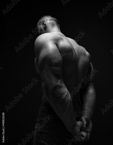 Strong muscular man, athlete, sportsman is standing naked, shirtless with his back to camera holding hands behind back and head bowed over dark background