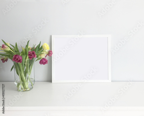 A model of a painting in a white frame in the interior on a table with a bouquet of pink and yellow tulips in a glass vase