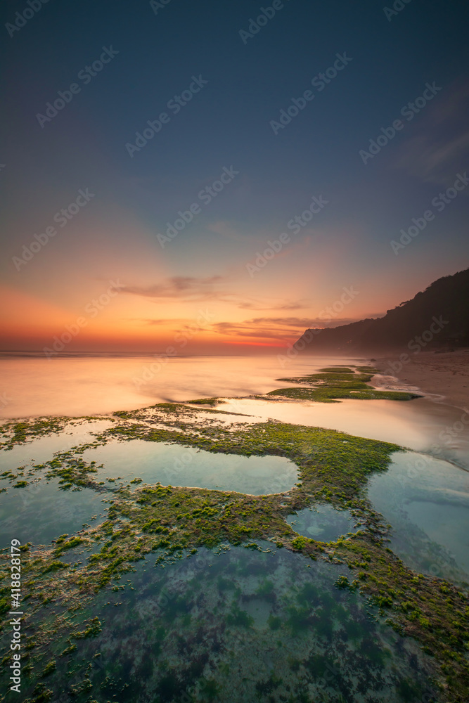Seascape for background. Sunset time. Beach with rocks and stones. Low tide. Stones with green seaweed and moss. Blue sky with sunlight on horizon. Copy space. Melasti beach, Bali
