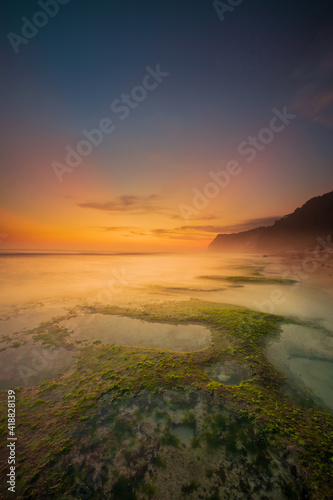 Seascape for background. Sunset time. Beach with rocks and stones. Low tide. Stones with green seaweed and moss. Blue sky with sunlight on horizon. Copy space. Melasti beach  Bali
