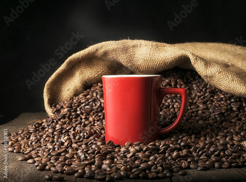 Red mug with hot coffee on the background of a heap of coffee beans spilled from a bag on the table