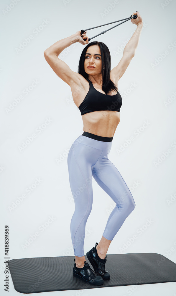 Attractive and muscular sportswoman dressed in stylish sportswear with elastic resistance bands poses in white background. Attractive and muscular sportswoman dressed in stylish sportswear with elastic resistance bands poses in white background.