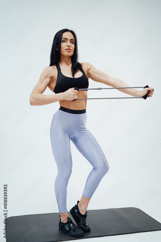 Attractive and muscular sportswoman dressed in stylish sportswear with elastic resistance bands poses in white background.