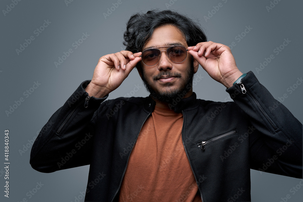 Obraz premium Stylish hindu hipster wearing fashionable clothing poses in gray background taking off his sunglasses.