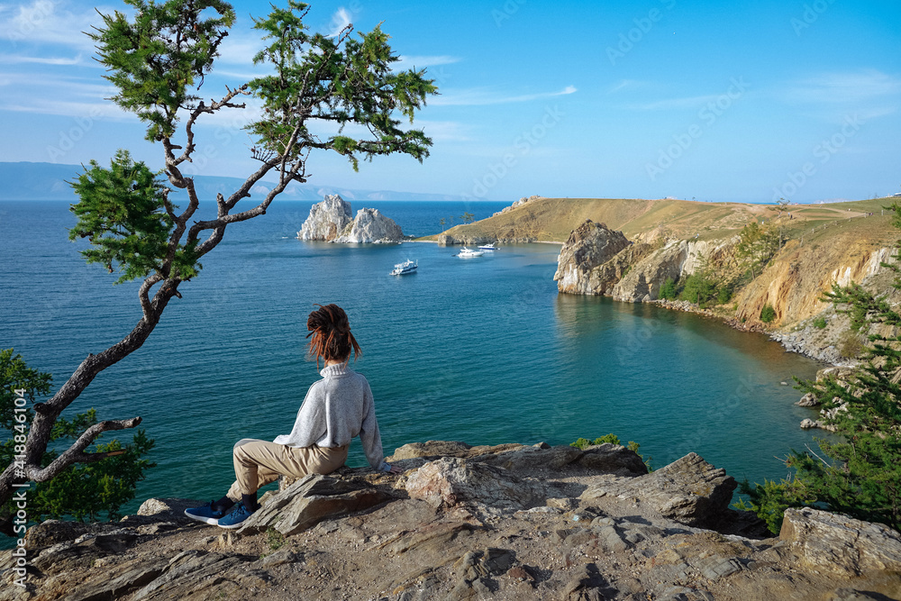 Girl with dreadlocks sits on rock under pine tree and looks at bay. Summer European view. Travel concept