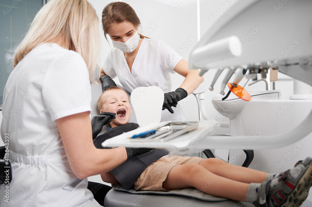 Little boy sitting in dental chair while two female dentists checking kid teeth. Dentist examining boy teeth with dental instrument. Concept of pediatric dentistry and dental care.