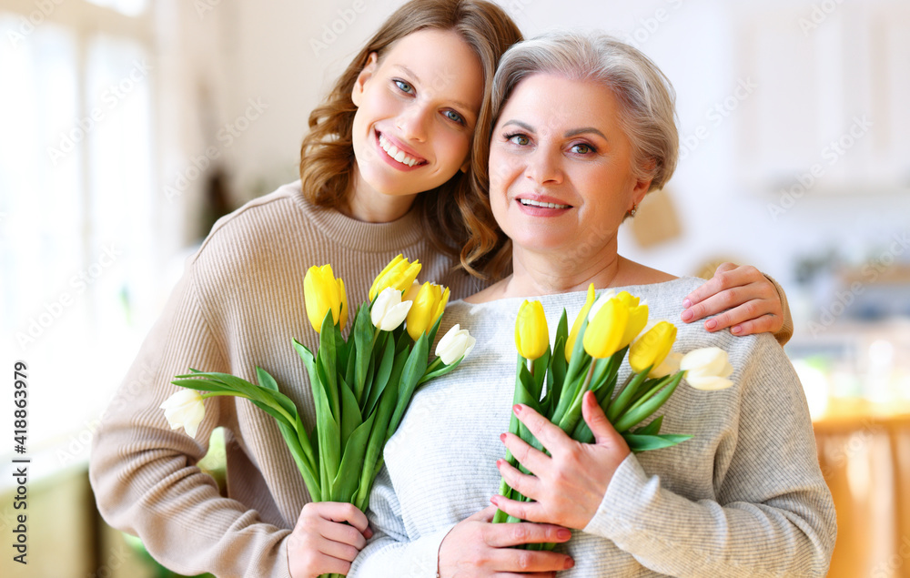 Happy lady embracing elderly mother while standing in apartment with tulips bouquets