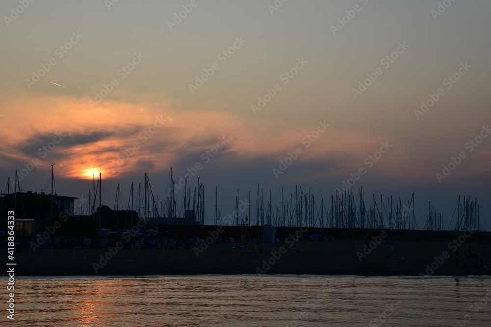 Romantic sunset over the harbor. Red-golden sunset in the background with masts of yachts and sailboats. Sunset on the Italian beach. Dark sea masts in the harbor and the sun in the clouds. End of day