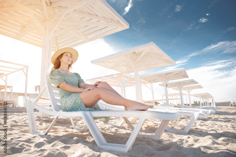 Young attractive caucasian woman in a green dress and a straw sits resting on a lounger under a wooden umbrella on a sandy beach of the seashore or ocean.