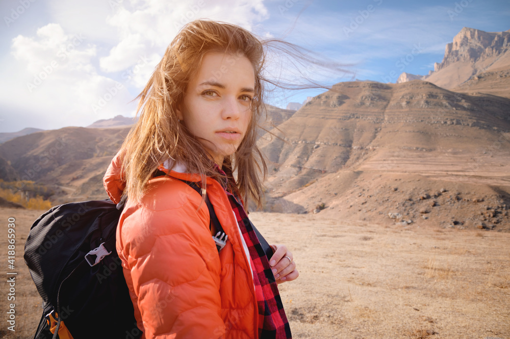 Young attractive Jewish woman in a down jacket and with a backpack against the backdrop of the epic mountains. Looks pensively at the camera
