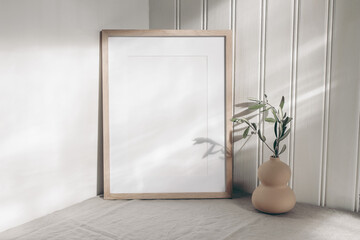 Portrait empty wooden frame mockup in sunlight. Olive branch in modern organic shaped vase. Beige linen table cloth. White wainscot wall paneling background. Scandinavian interior, home design. Art