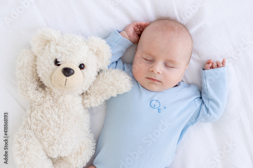 baby boy sleeps on the bed lying on his back with a soft toy bear in blue pajamas holding his hands up, healthy newborn sleep