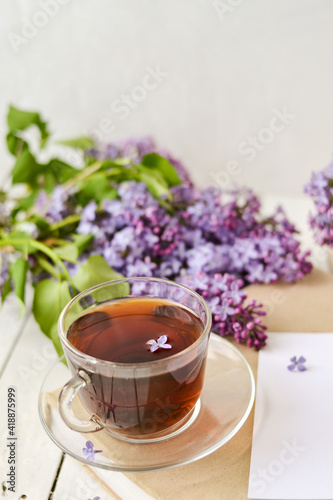 Romantic background with a cup of tea, lilac flowers and a book over a white wooden table. Leisure concept, spring breakfast