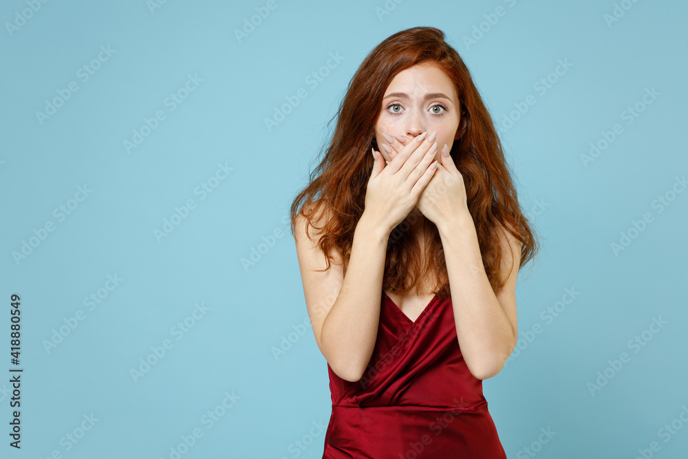Young caucasian scared frightened shocked surprised readhead confused curly woman in red party evening dress gown cover mouth with hands omg gesture isolated on pastel blue background studio portrait. Young caucasian scared frightened shocked surprised readhead confused curly woman in red party evening dress gown cover mouth with hands omg gesture isolated on pastel blue background studio portrait.