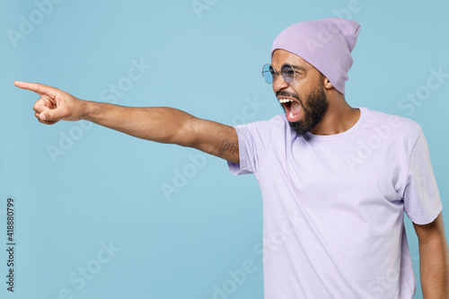 Young angry indignant strict unshaven student black african man 20s inviolet t-shirt hat glasses point index finger aside command do it scream shout isolated on pastel blue background studio portrait.