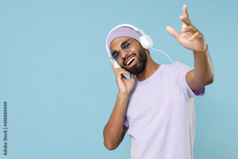 Young cool unshaven black african man 20s wear violet t-shirt hat glasses white headphones listen to music stretching hand to camera dancing isolated on pastel blue color background studio portrait Young cool unshaven black african man 20s wear violet t-shirt hat glasses white headphones listen to music stretching hand to camera dancing isolated on pastel blue color background studio portrait