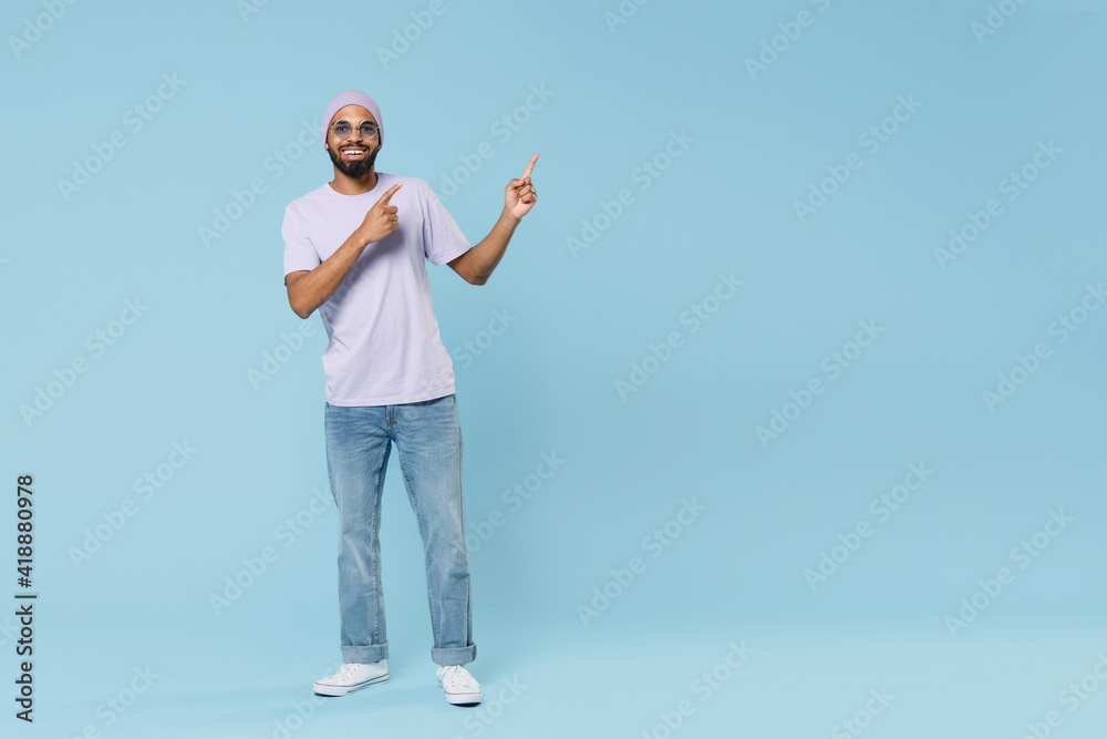 Full length young cool smiling happy fun unshaven black african man 20s wear violet t-shirt hat glasses point index finger aside on workspace area isolated on pastel blue background studio portrait