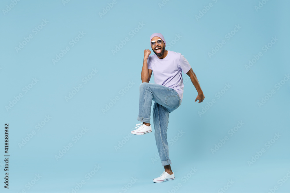 Full length young happy fun unshaven black african man in violet t-shirt hat glasses do winner gesture with raised leg clench fist celebrate isolated on pastel blue color background studio portrait Full length young happy fun unshaven black african man in violet t-shirt hat glasses do winner gesture with raised leg clench fist celebrate isolated on pastel blue color background studio portrait