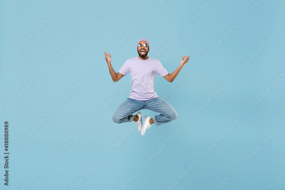 Full length young unshaven black african man in violet t-shirt hat glasses hold spread hands in yoga om gesture relax meditate jump high levitate isolated on pastel blue background studio portrait.