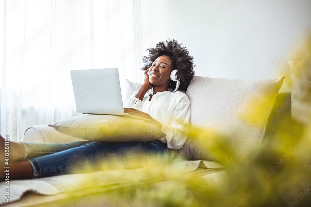 Relaxed young African woman with eyes closed sitting on her bed enjoying music over headphones from smartphone at home. Shot of a young woman using a laptop and headphones on the sofa at home
