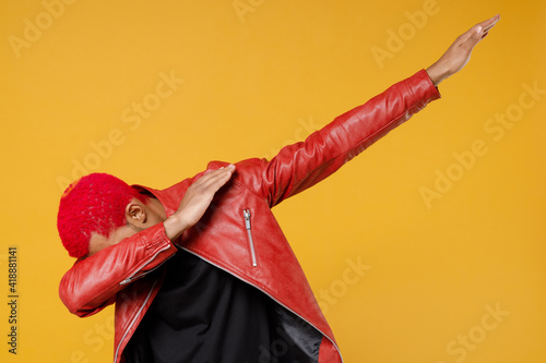 Young african man with funky trendy pink hairdo in red leather jacket doing dab hip hop dance hands move gesture youth sign hiding covering face isolated on yellow orange background studio portrait.