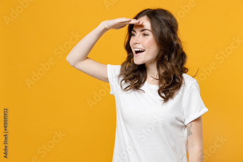 Young happy smiling excited fun caucasian student woman wearing white basic casual t-shirt hold hand at forehead looking far away distance isolated on yellow orange color background studio portrait