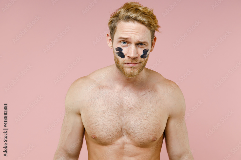 Fototapeta premium Confused bearded naked young man 20s years old perfect skin smear clay facial mask on cheek isolated on pastel pink color background studio portrait. Skin care healthcare cosmetic procedures concept.