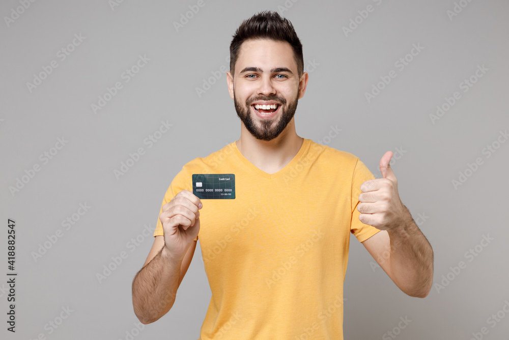 Young caucasian smiling bearded attractive rich successful happy man 20s in casual yellow basic t-shirt hold credit bank card show thumb up gesture isolated on grey color background studio portrait. Young caucasian smiling bearded attractive rich successful happy man 20s in casual yellow basic t-shirt hold credit bank card show thumb up gesture isolated on grey color background studio portrait.