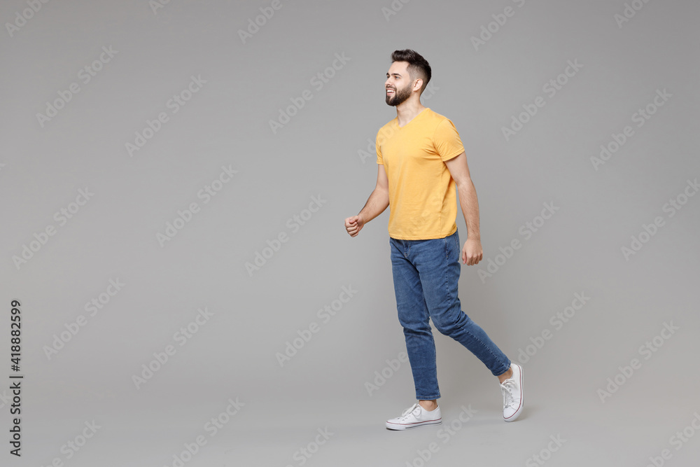 Full length side profile view young caucasian serious bearded attractive handsome student man 20s wear casual yellow basic t-shirt walking going isolated on grey color background studio portrait . Full length side profile view young caucasian serious bearded attractive handsome student man 20s wear casual yellow basic t-shirt walking going isolated on grey color background studio portrait .