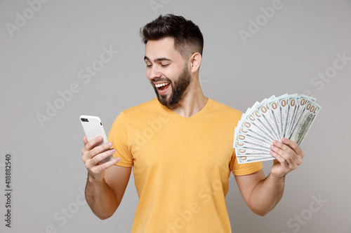 Obraz na plátně Young bearded rich happy man 20s wear casual yellow basic t-shirt holding mobile cell phone holding fan of cash money in dollar banknotes shopping online isolated on grey background studio portrait