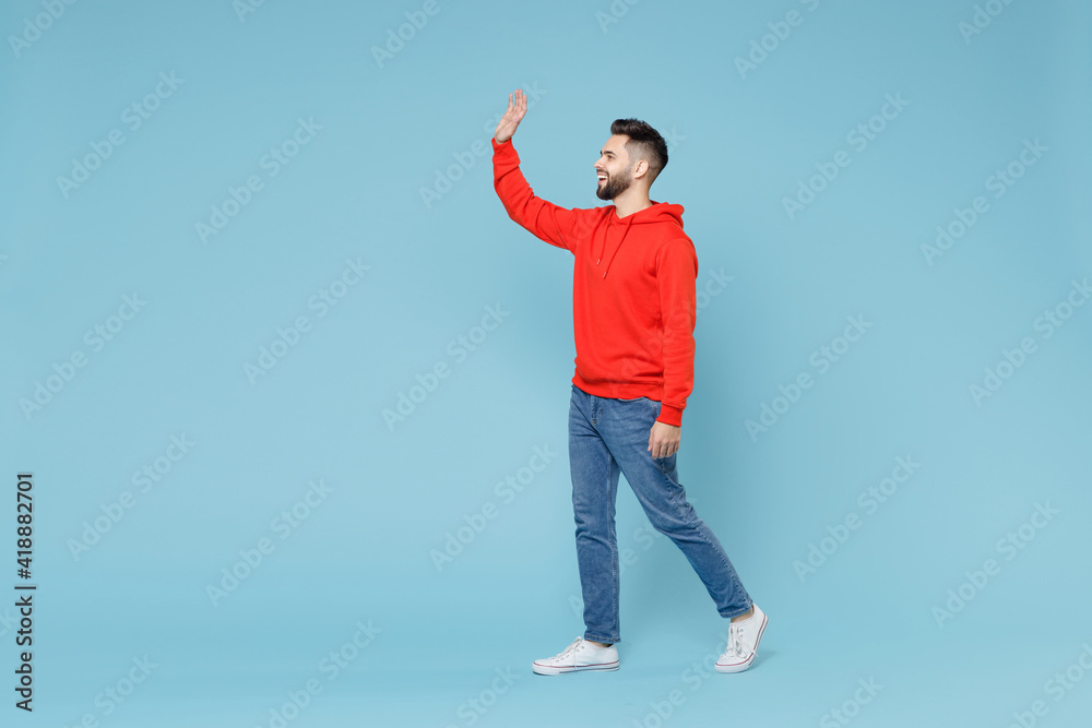 Full length side profile view of young caucasian smiling bearded man 20s in casual red orange hoodie waving hand greeting someone isolated on blue background studio portrait People lifestyle concept. Full length side profile view of young caucasian smiling bearded man 20s in casual red orange hoodie waving hand greeting someone isolated on blue background studio portrait People lifestyle concept.