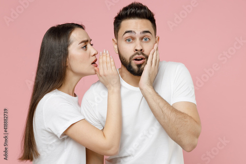 Young couple two friends bearded man brunette woman in white basic blank print design t-shirt whispering gossip and tells secret behind her hand, sharing news isolated on pastel pink color background
