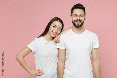 Young cheerful couple two friends bearded man brunette woman in white basic blank print design t-shirts posing looking camera standing smiling isolated on pastel pink color background studio portrait.