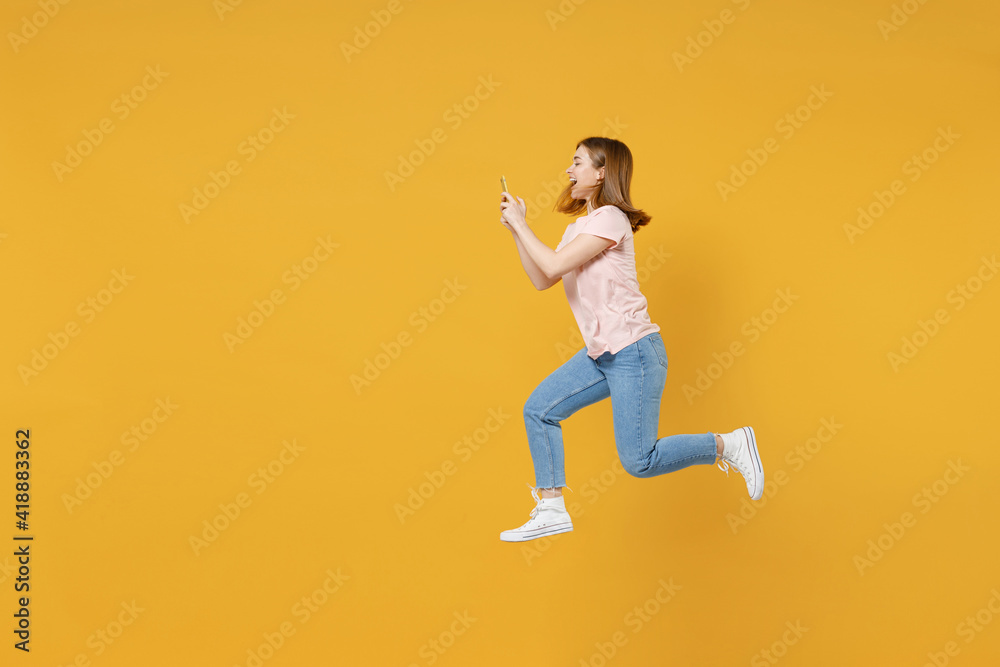 Full length side profeli view of young expressive excited overjoyed woman 20s in basic pastel pink t-shirt jump high run holding mobile cell phone using fast internet isolated on yellow background .