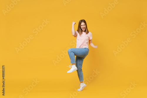 Full length of young caucasian overjoyed excited woman 20s in basic pastel pink t-shirt, jeans do winner gesture clench fist celebrating, raised up leg isolated on yellow background studio portrait.